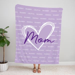 Może przedstawiać: Fioletowy koc z białym sercem i napisem "Mama" w kolorze fioletowym. Koc jest pokryty powtarzającym się wzorem imion, w tym "Emma", "Harry", "Olivia" i "Marry".