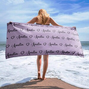 May include: A lavender beach towel with the name "Amelia" repeated in a black cursive font with small hearts in between each name.