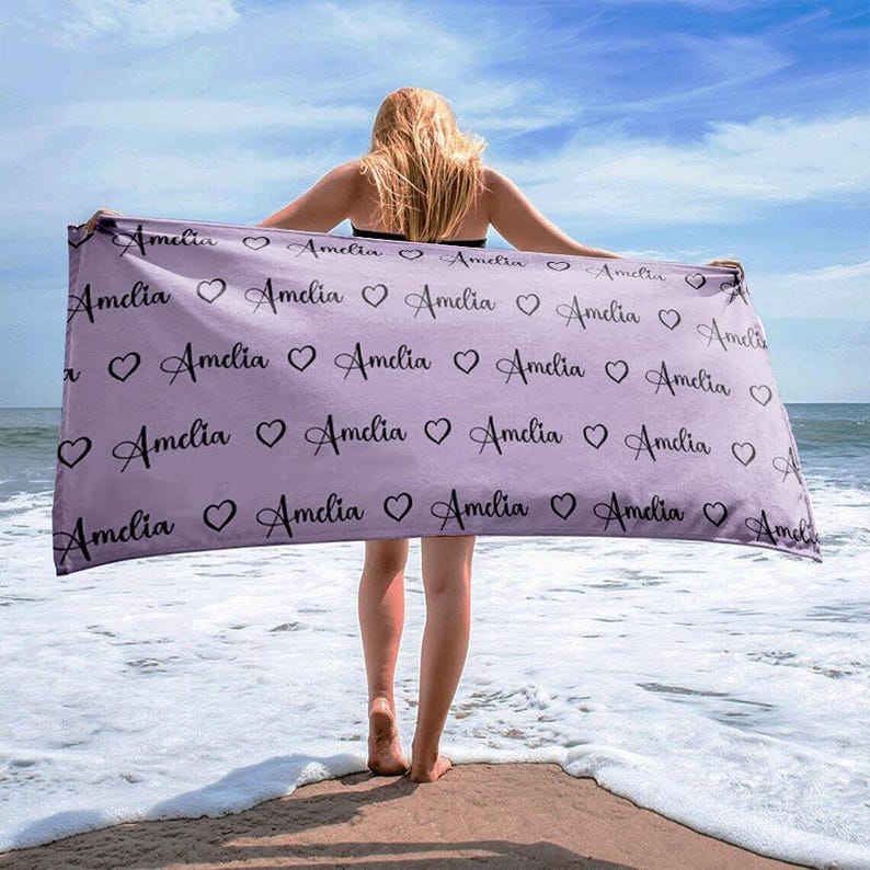 May include: A lavender beach towel with the name "Amelia" repeated in a black cursive font with small hearts in between each name.