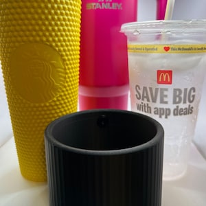 Puede incluir: Un portavasos cilíndrico negro con nervaduras se encuentra delante de tres recipientes de bebidas. Los recipientes son una taza de Starbucks amarilla y verde, un vaso Stanley rosa y un vaso de plástico transparente con el logotipo de McDonald's y el texto "Save Big with app deals".