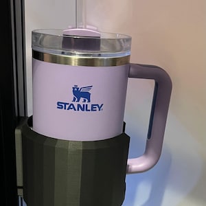 May include: A purple stainless steel travel mug with a black handle and a black and gray cup holder. The mug has a clear lid and a straw. The mug has the Stanley logo on the side.