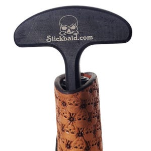 May include: A black bottle opener with a skull logo and the text "Slickbald.com" on the handle. The handle is connected to a brown leather grip with a repeating skull pattern.