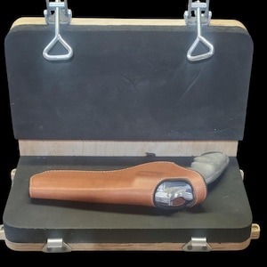 May include: A handgun in a brown leather holster rests inside an open wooden case with a black interior. The case has metal latches and hinges. The holster is light brown with white stitching.