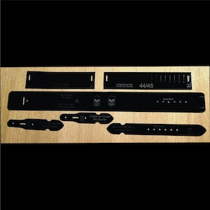 May include: Black leather belt making templates with various shapes and sizes. The templates include text such as "36/38", "44/45", and "Shape for Center Bar Buckle". The templates are arranged on a wooden surface.