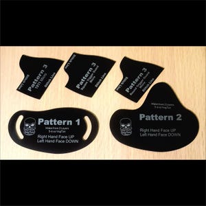 May include: Black leathercraft patterns with white text on a light wood surface. The patterns include shapes for a trigger guard and face mask, with text indicating layers and veg tan weight. A skull graphic is also present.