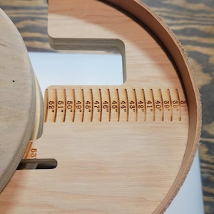 May include: A light-colored wooden belt sizing tool with a circular base and a curved edge. The tool features laser-engraved markings ranging from 37 inches to 53 inches. The text "Hygiene Colorado USA Slickbald Customs" is visible on the surface.