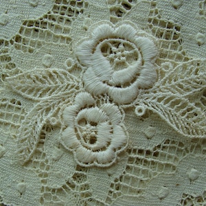 May include: Close-up of delicate white lace fabric with intricate floral designs. The lace features two large flowers with detailed stitching and smaller leaves.