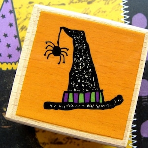 May include: A wooden stamp with an orange background features a black witch's hat with a spider hanging from it. The hat has a purple and green band. The stamp is a Halloween-themed craft supply.