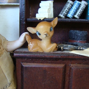 May include: A small, brown and white ceramic deer figurine with blue eyes. The figurine is sitting on a wooden shelf with books and a hat.