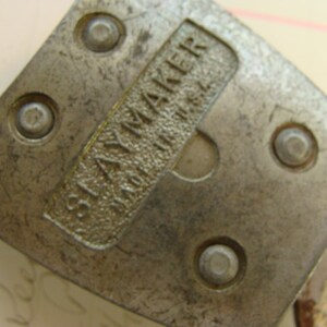 May include: Close-up of a silver metal tool with the text "SLAYMAKER" and a series of five small, round holes.