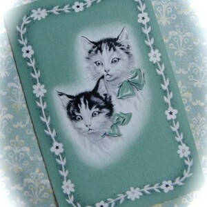 May include: A vintage greeting card with a green background and a white border. The border is decorated with white flowers. Two black and white kittens are featured on the card. The kittens are looking at the viewer. The card has a vintage feel.