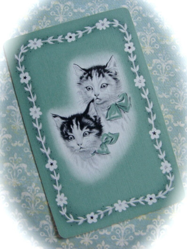 May include: A vintage greeting card with a green background and a white border. The border is decorated with white flowers. Two black and white kittens are featured on the card. The kittens are looking at the viewer. The card has a vintage feel.
