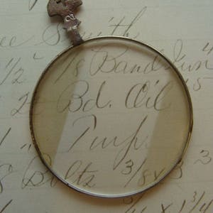 May include: A vintage magnifying glass with a silver frame and a small key-shaped handle. The glass is round and has a clear lens. The key has a cross on it. The glass is resting on a piece of paper with handwritten text that reads "1/8 Band, 1/2 Bel. Oil,  Tynj,  3/8, 1/16 x 1 1/4".