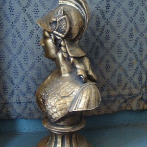 May include: A gold-colored bust sculpture of a woman wearing a helmet with a feathered plume. The bust is on a pedestal with a fluted base.