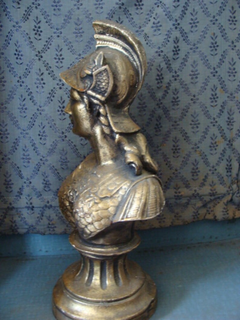 May include: A gold-colored bust sculpture of a woman wearing a helmet with a feathered plume. The bust is on a pedestal with a fluted base.