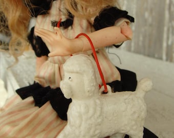 May include: A doll with blonde hair wearing a pink and white striped dress with a black lace trim and a red ribbon holding a white toy poodle.