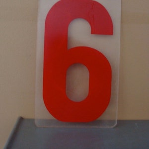 May include: A red number six on a clear plastic rectangle. The number is displayed on a light gray shelf. The background is a light beige wall.