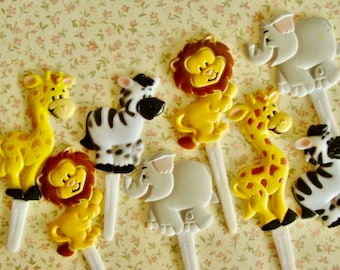 May include: A collection of colorful plastic animal figurines on white sticks. The set includes giraffes, zebras, lions, and elephants in yellow, white, and gray. The figurines are arranged on a floral patterned background.