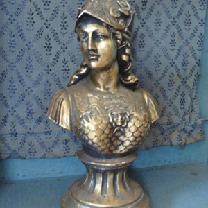 May include: A gold-colored bust sculpture of a woman wearing a helmet. The bust is on a pedestal with a round base.