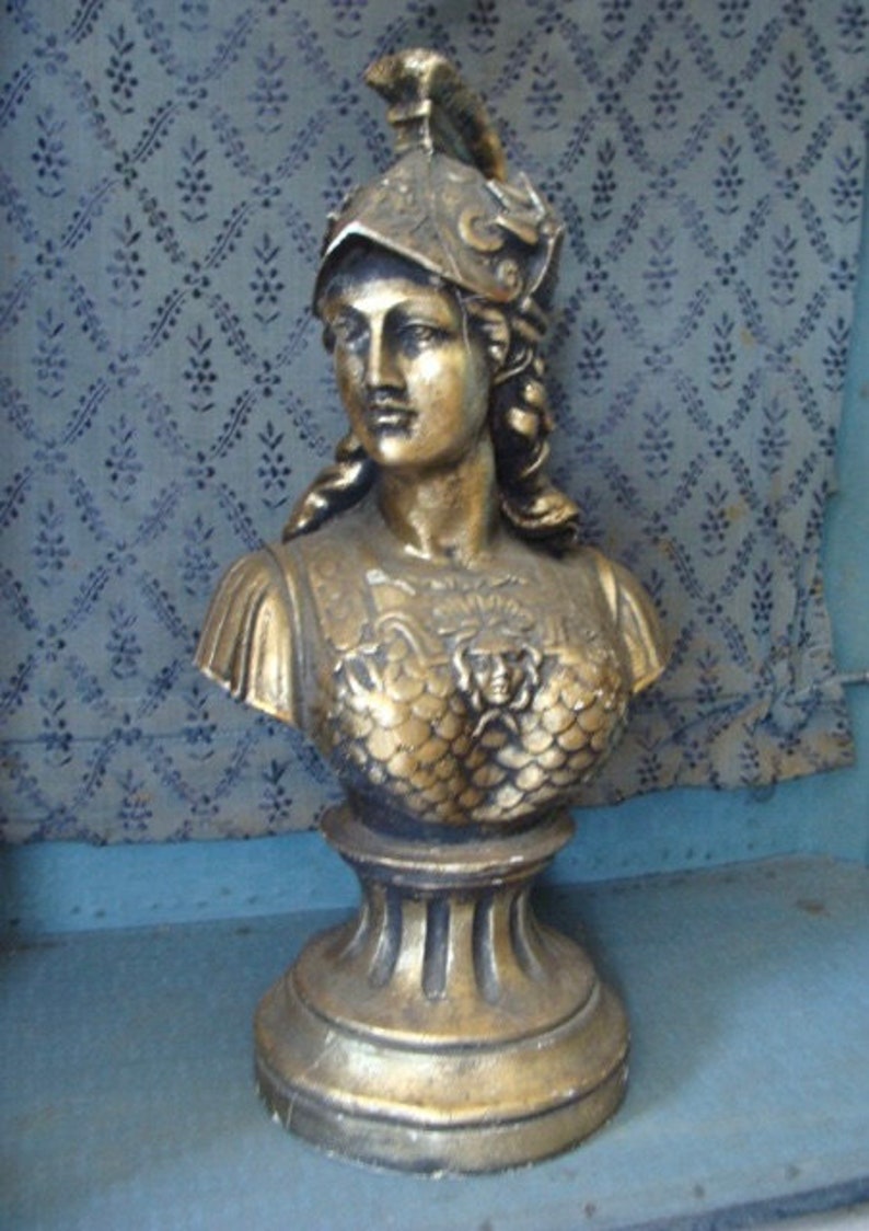 May include: A gold-colored bust sculpture of a woman wearing a helmet. The bust is on a pedestal with a round base.