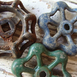 May include: Three vintage metal water valve handles in different colors: rusty brown, blue, and green. Each handle has a unique design with multiple points.