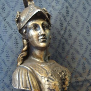 May include: A gold-colored bust sculpture of a woman wearing a helmet with a decorative design. The bust is detailed with a textured, scaly pattern on the chest and a flowing fabric draped over the shoulders.