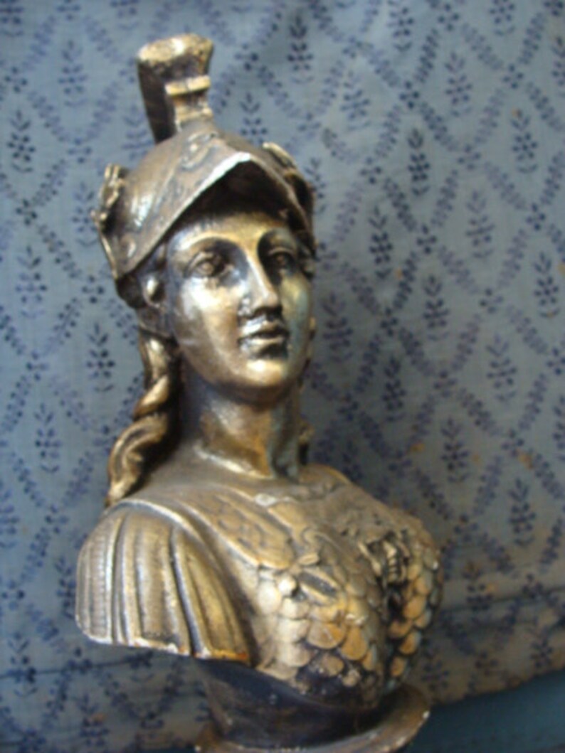 May include: A gold-colored bust sculpture of a woman wearing a helmet with a decorative design. The bust is detailed with a textured, scaly pattern on the chest and a flowing fabric draped over the shoulders.