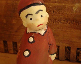 Może przedstawiać: Mała ceramiczna figurka przedstawiająca osobę w czerwonym kapeluszu i płaszczu z białymi guzikami. Figurka ma namalowaną twarz z poważnym wyrazem.