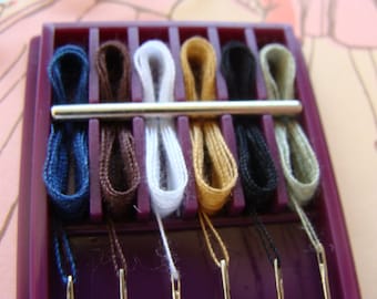 May include: A purple plastic sewing kit with eight spools of thread in different colors, including blue, brown, white, gold, black, and beige. The spools are arranged in a row and have a metal bar across the top.