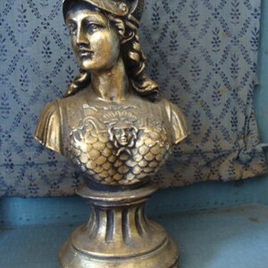 May include: A gold-colored bust sculpture of a woman wearing a helmet with a decorative design. The bust is on a pedestal with a fluted base.