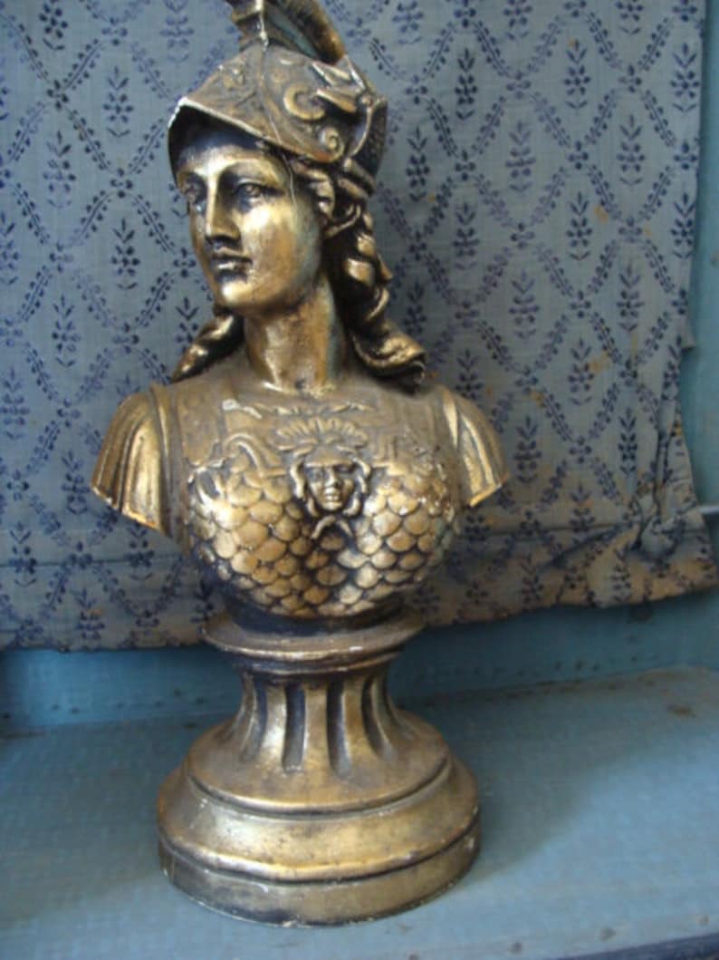 May include: A gold-colored bust sculpture of a woman wearing a helmet with a decorative design. The bust is on a pedestal with a fluted base.