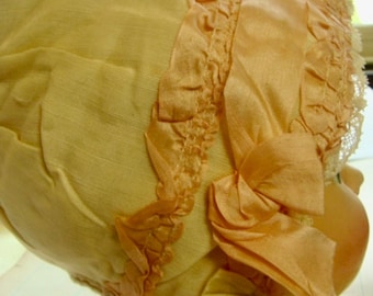 May include: A vintage baby bonnet made of white fabric with a pink satin ribbon and lace trim.