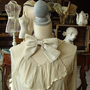 Puede incluir: Un maniquí blanco con un gran lazo alrededor del cuello. El maniquí lleva un vestido blanco con ribete de encaje. El maniquí está de pie sobre una mesa de madera con un mantel blanco.