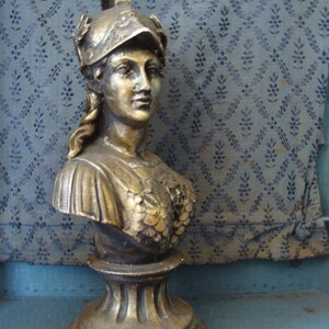 May include: A gold-colored bust sculpture of a woman wearing a helmet. The bust is on a pedestal with a decorative base.