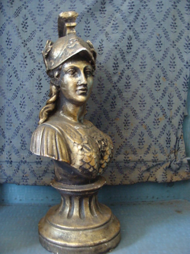 May include: A gold-colored bust sculpture of a woman wearing a helmet. The bust is on a pedestal with a decorative base.