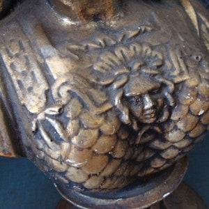 May include: A gold-painted plaster bust of a woman with a detailed, textured design. The bust is wearing a decorative, ornate garment with a sculpted face of a woman with a crown.