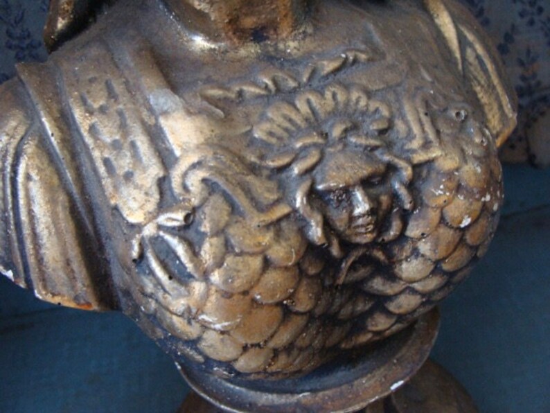 May include: A gold-painted plaster bust of a woman with a detailed, textured design. The bust is wearing a decorative, ornate garment with a sculpted face of a woman with a crown.