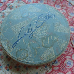 May include: A blue and white round tin with the text "Lady Esther" printed on it. The tin has a floral design and is likely for cosmetics.