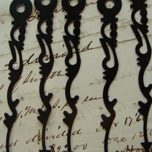 May include: Five black metal clock hands with ornate designs. The hands are all the same size and shape, and they have a pointed tip. The hands are arranged in a row, and they are all pointing in the same direction.