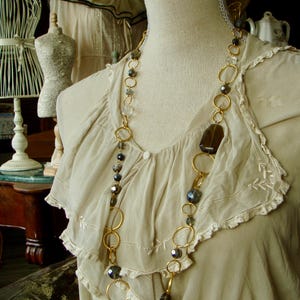 Puede incluir: Un collar de cadena dorada con cuentas de vidrio gris oscuro y transparentes y un gran colgante de piedra marrón. El collar se muestra en un maniquí blanco con un ribete de encaje blanco.