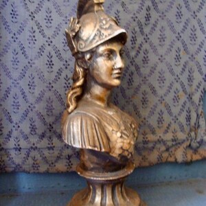 May include: A gold-painted bust sculpture of a woman wearing a helmet with intricate details. The sculpture is on a pedestal with a fluted design.
