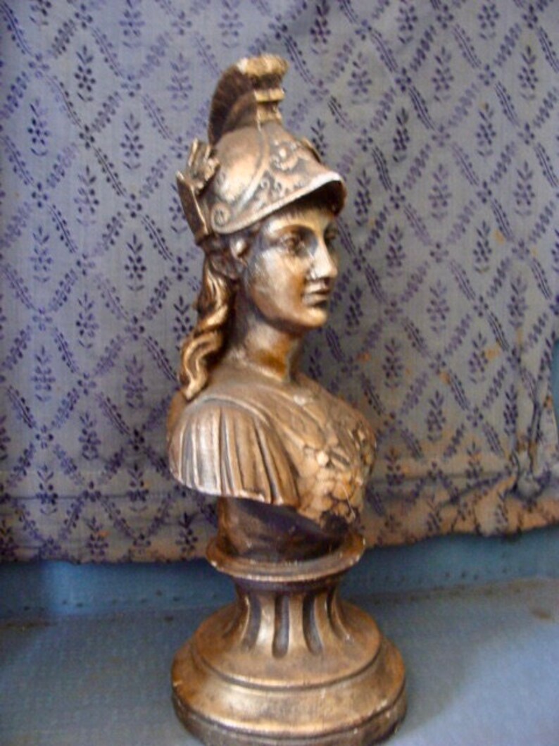 May include: A gold-painted bust sculpture of a woman wearing a helmet with intricate details. The sculpture is on a pedestal with a fluted design.