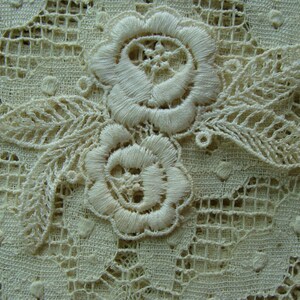 May include: Close-up of delicate white lace fabric with a floral design. The lace features two embroidered flowers with intricate details and leaves.