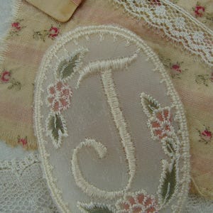 May include: Oval-shaped embroidered patch with the letter "J" in cream-colored thread. The patch is adorned with pink floral and green leaf accents. A spool of pink thread and a floral fabric are in the background.