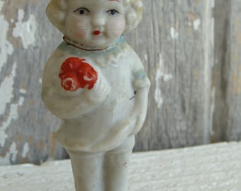 Pode incluir: Uma figura de boneca de porcelana de uma menina com cabelo loiro, vestindo um vestido branco com detalhes vermelhos e guarnições azuis. Ela está segurando um buquê de flores vermelhas em suas mãos.