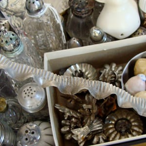 May include: A collection of vintage glass salt and pepper shakers in a box with various metal trinkets. The shakers are clear glass with silver tops and some have a textured surface. The box is white with a brown interior.