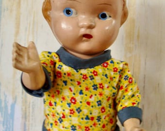 May include: Vintage doll with a painted face, blue eyes, and a yellow floral dress with blue trim. The doll's hair is brown, and it has its arm raised. The doll is a classic toy.