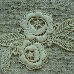 May include: White lace embroidery with two floral designs and leaves on a beige fabric background.