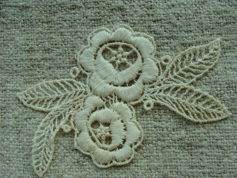 May include: White lace embroidery with two floral designs and leaves on a beige fabric background.
