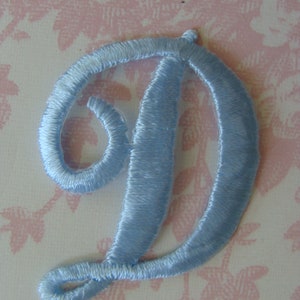 May include: A light blue embroidered letter "D" patch. The script letter is made of a satin-like thread and is stitched onto a pink floral patterned background. The patch is a decorative element for clothing or crafts.